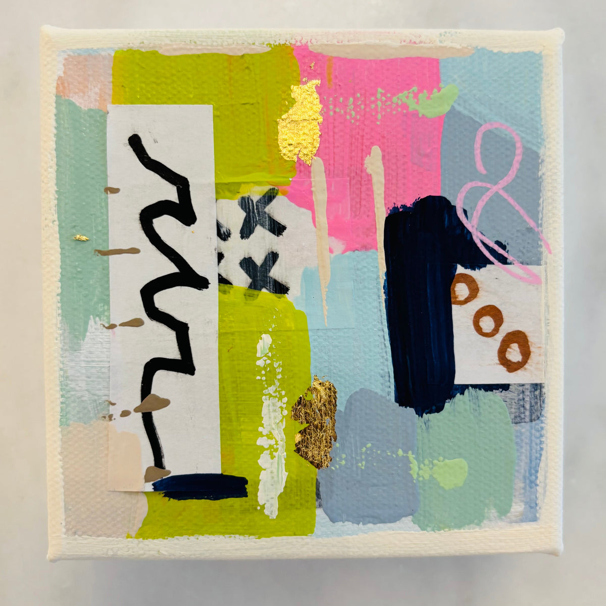 A square abstract canvas painting with textured brushstrokes in pastel pink, light blue, mint green, olive yellow, black, and white, featuring gold leaf accents, displayed on a light marble surface.