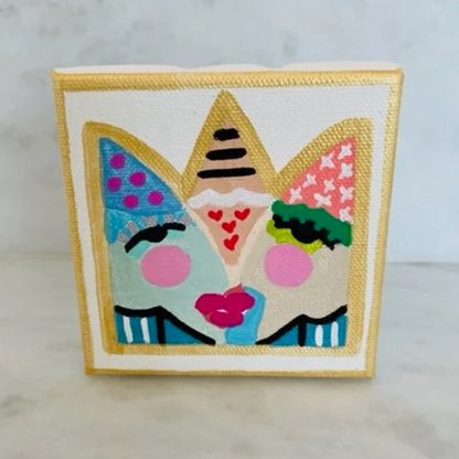 A square canvas art print featuring a whimsical, colorful illustration of three faces wearing party hats, displayed on a light marble surface against a light gray marble wall.
