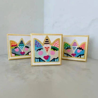 Three small embroidered patch-style art pieces with colorful abstract faces and gold borders, arranged on a light marble surface against a pale wall.