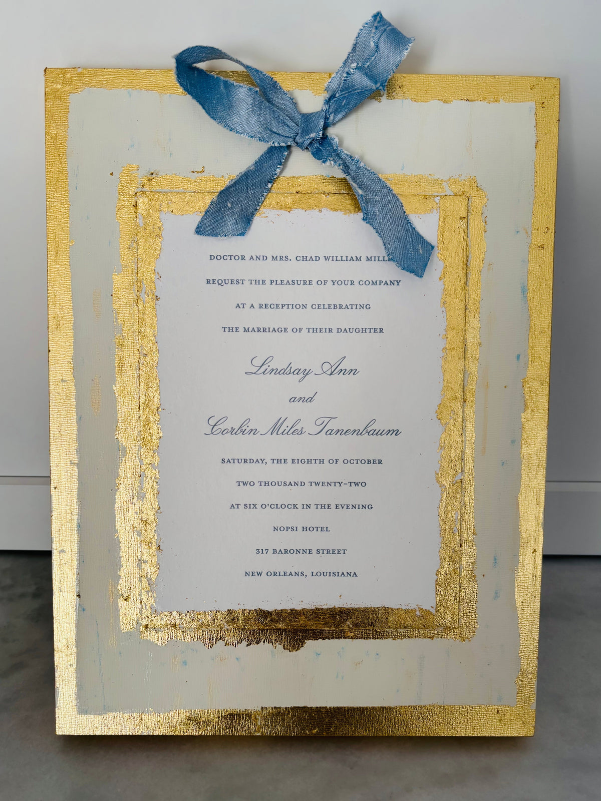 A square invitation card with a distressed gold frame, a white center panel, and a blue ribbon tied at the top, photographed against a light gray wall and gray surface.