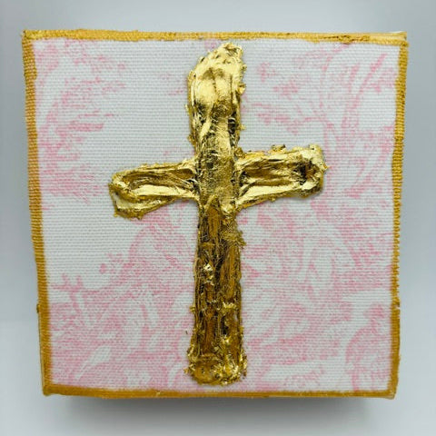 A square textile piece featuring a raised metallic gold cross on a white background with a light pink floral pattern, finished with a gold border.