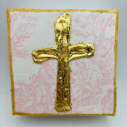 A square textile piece featuring a raised metallic gold cross on a white background with a light pink floral pattern, finished with a gold border.