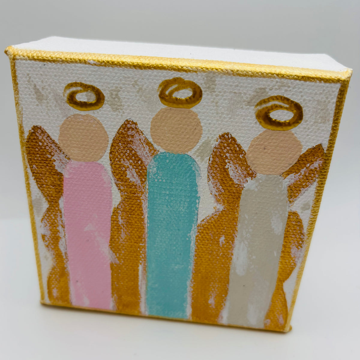 A small rectangular canvas painting showing three stylized angel figures with gold halos and gold wings, painted on a white textured background. The figures wear pink, aqua blue, and off-white robes.