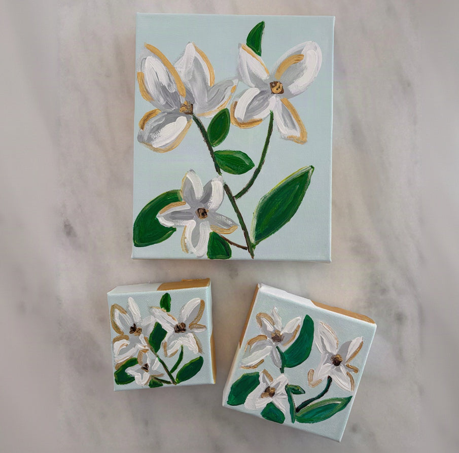 A set of three hand-painted canvas art pieces featuring white flowers with gold accents and green leaves on a pale blue background, arranged on a light marble surface.