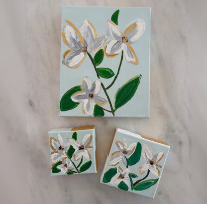 A set of three hand-painted canvas art pieces featuring white flowers with gold accents and green leaves on a pale blue background, arranged on a light marble surface.