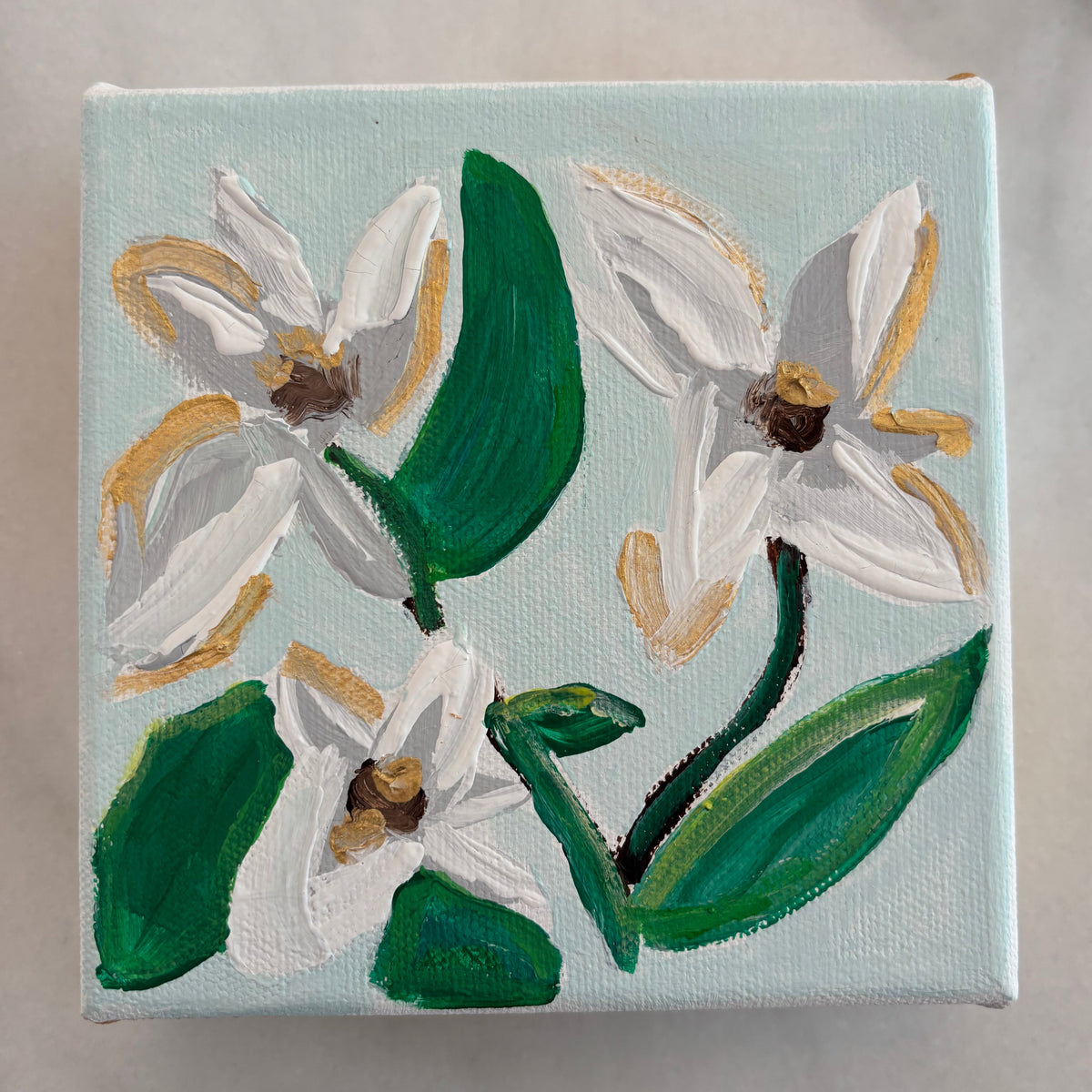 A square canvas painting featuring three white flowers with green leaves and gold accents on a pale blue background, displayed on a light gray marble surface.