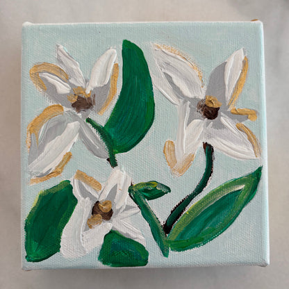 A square canvas painting featuring three white flowers with green leaves and gold accents on a pale blue background, displayed on a light gray marble surface.