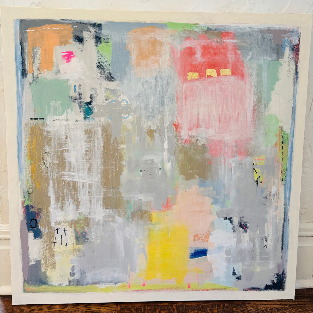 An abstract canvas painting with a white frame, featuring textured brushstrokes of gray, pink, yellow, green, and orange, leaning against a white textured wall.