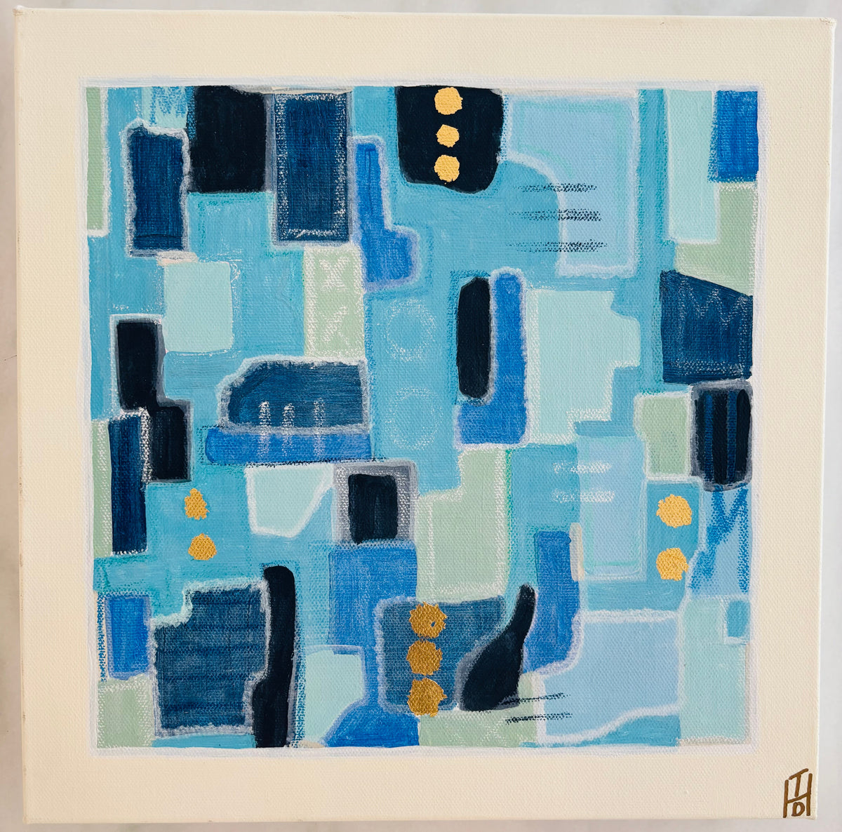 An abstract square painting featuring geometric blocks and shapes in various shades of blue, navy, light blue, and mint green, with small gold dots. The artwork is presented on a wide cream-colored mat board, photographed against a light gray marble surface.
