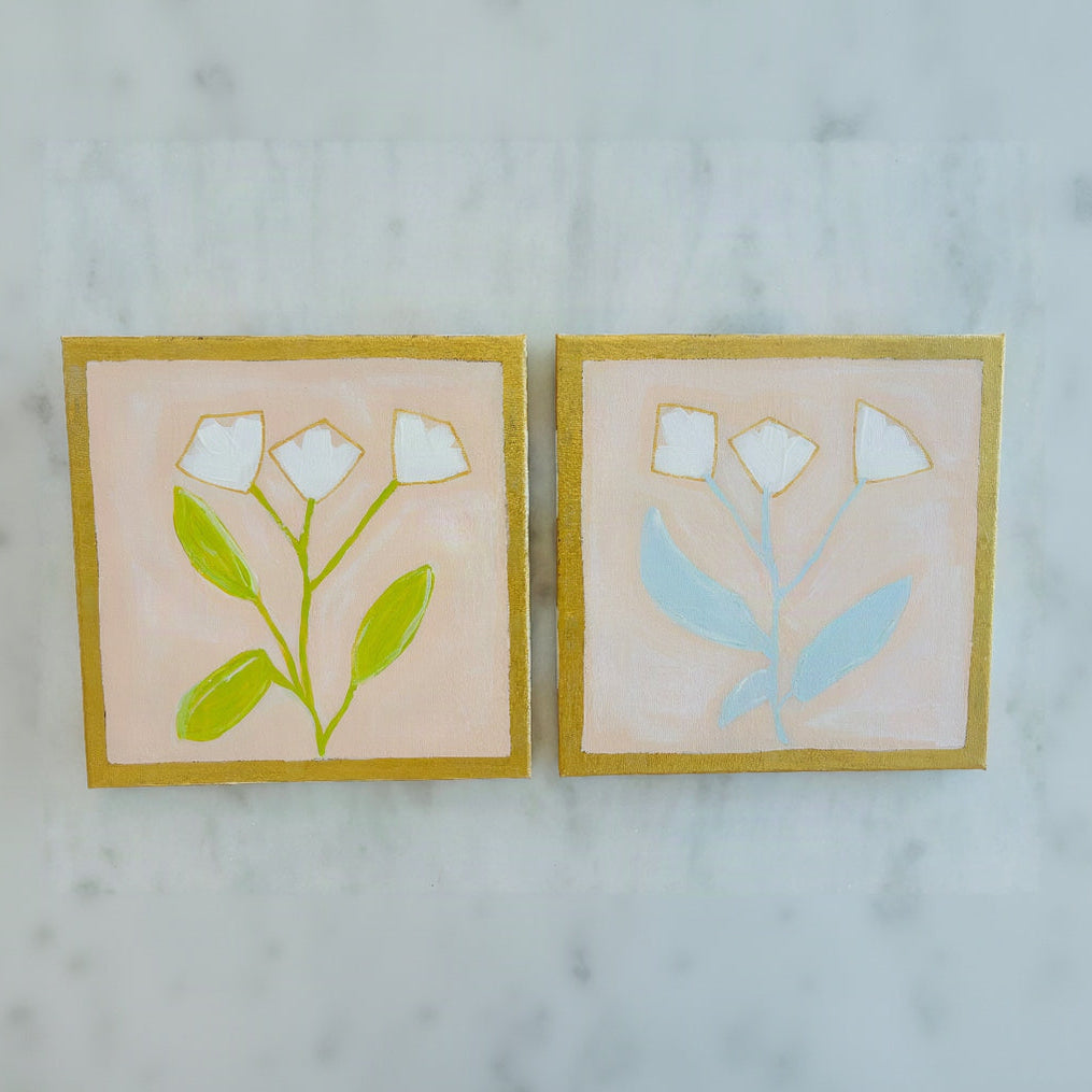 Two square hand-painted ceramic tiles featuring minimalist white flowers and green or light blue leaves on a soft pink background, framed by gold borders, displayed side-by-side on a light marble surface.