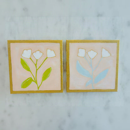 Two square hand-painted ceramic tiles featuring minimalist white flowers and green or light blue leaves on a soft pink background, framed by gold borders, displayed side-by-side on a light marble surface.