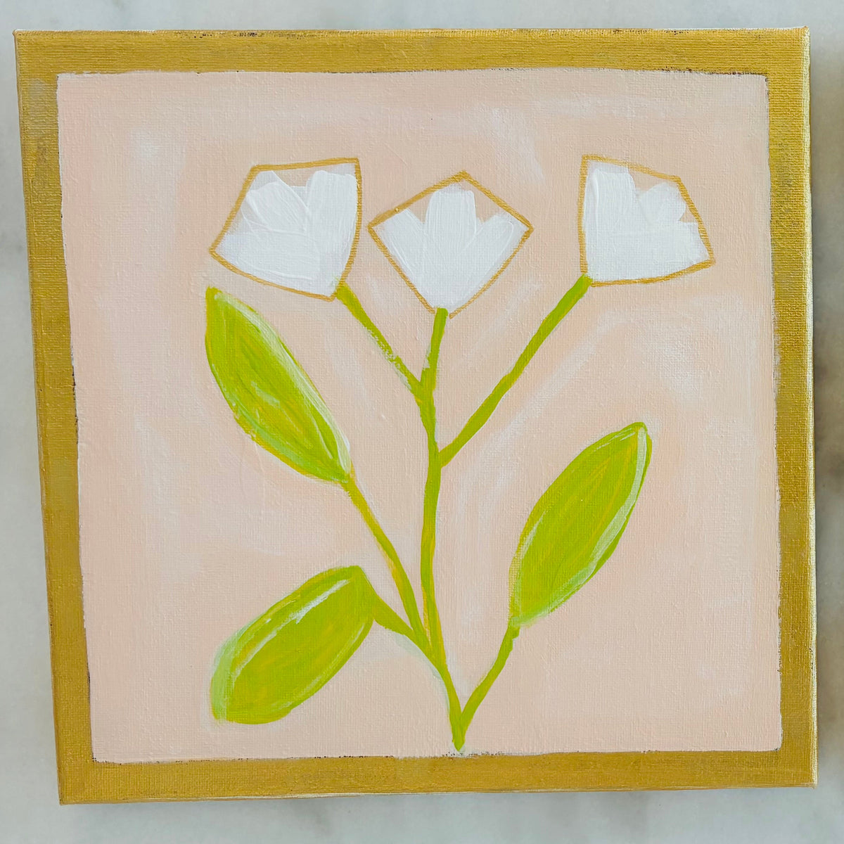 Two square hand-painted ceramic tiles featuring minimalist floral designs, displayed side-by-side on a light gray marble surface. The left tile depicts white flowers with green leaves on a soft pink background with a gold border.