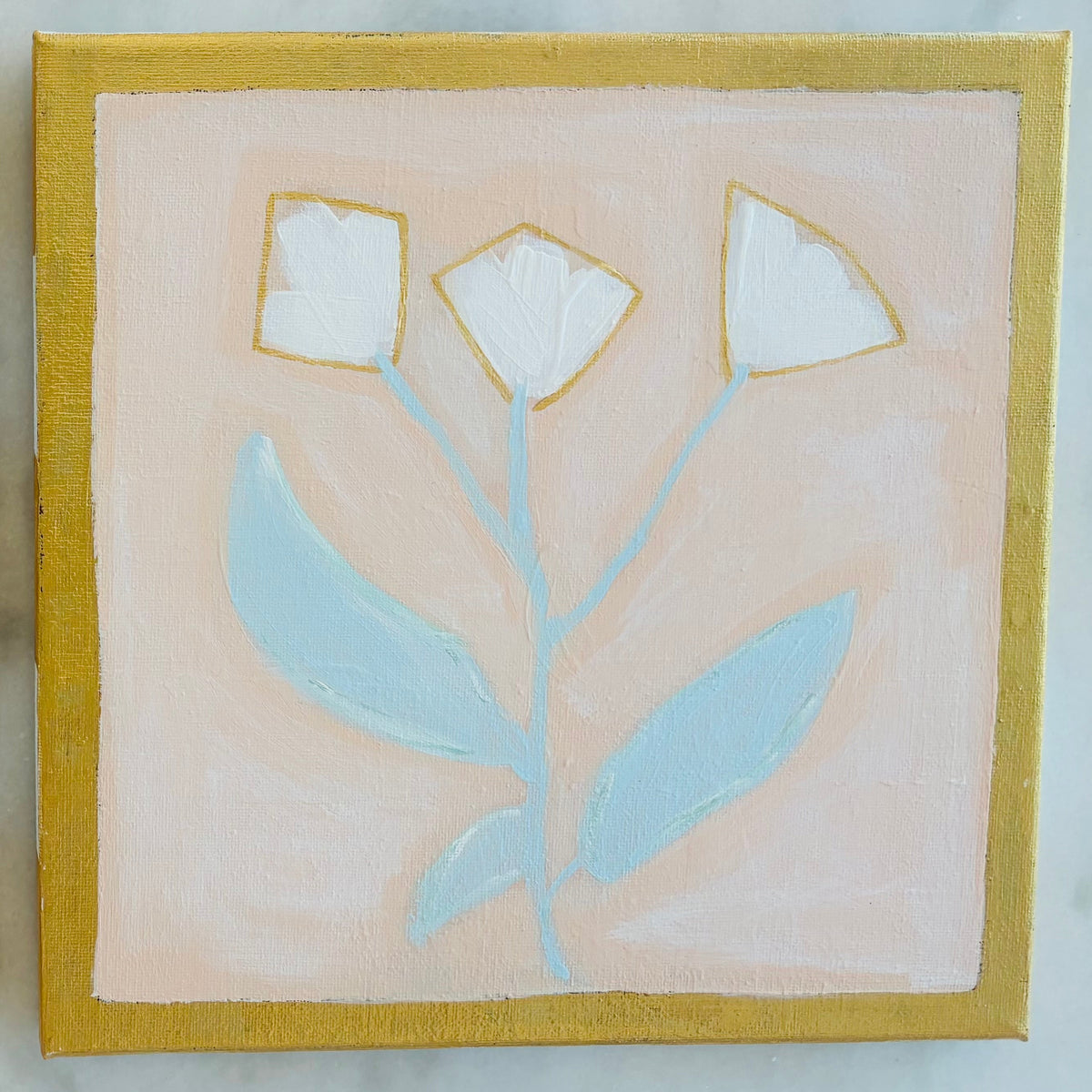 Two square hand-painted ceramic tiles featuring minimalist floral designs, displayed side-by-side on a light gray marble surface. The left tile depicts white flowers with green leaves on a soft pink background with a gold border.