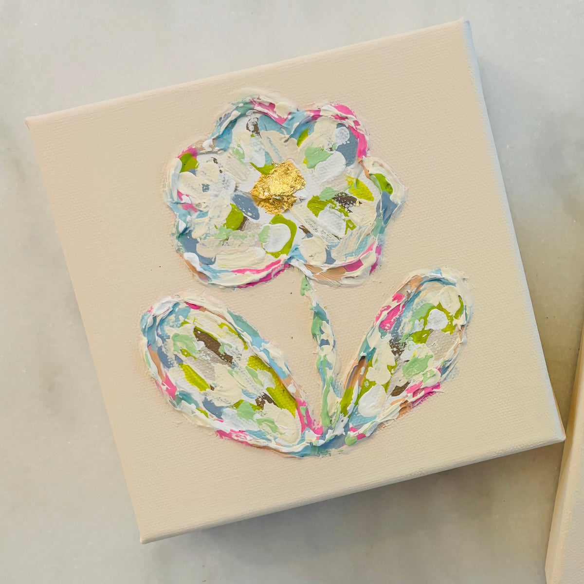 Two square cream-colored canvas art pieces featuring a stylized flower design made from layered, textured paper in pastel pink, green, blue, and white, with a small gold foil flower center, displayed on a light marble surface.
