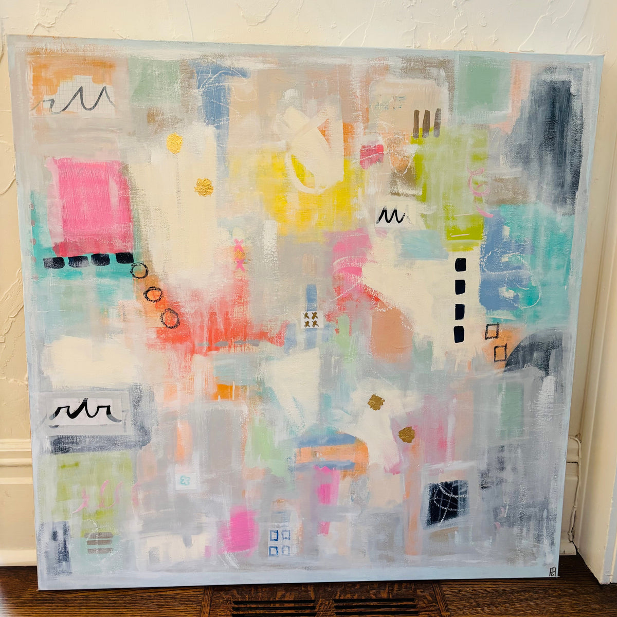 An abstract canvas painting with a light blue-gray border, featuring a collage of pastel and bright colors including pink, yellow, teal, orange, and white, leaning against a textured off-white wall on a wooden floor.