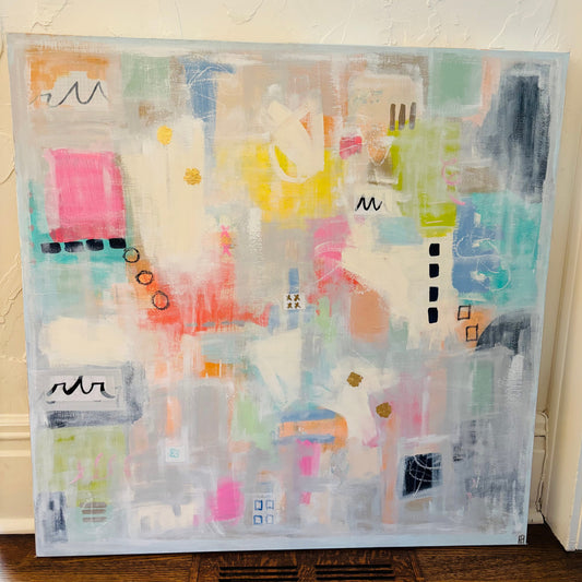 An abstract canvas painting with a light blue-gray border, featuring a collage of pastel and bright colors including pink, yellow, teal, orange, and white, leaning against a textured off-white wall on a wooden floor.